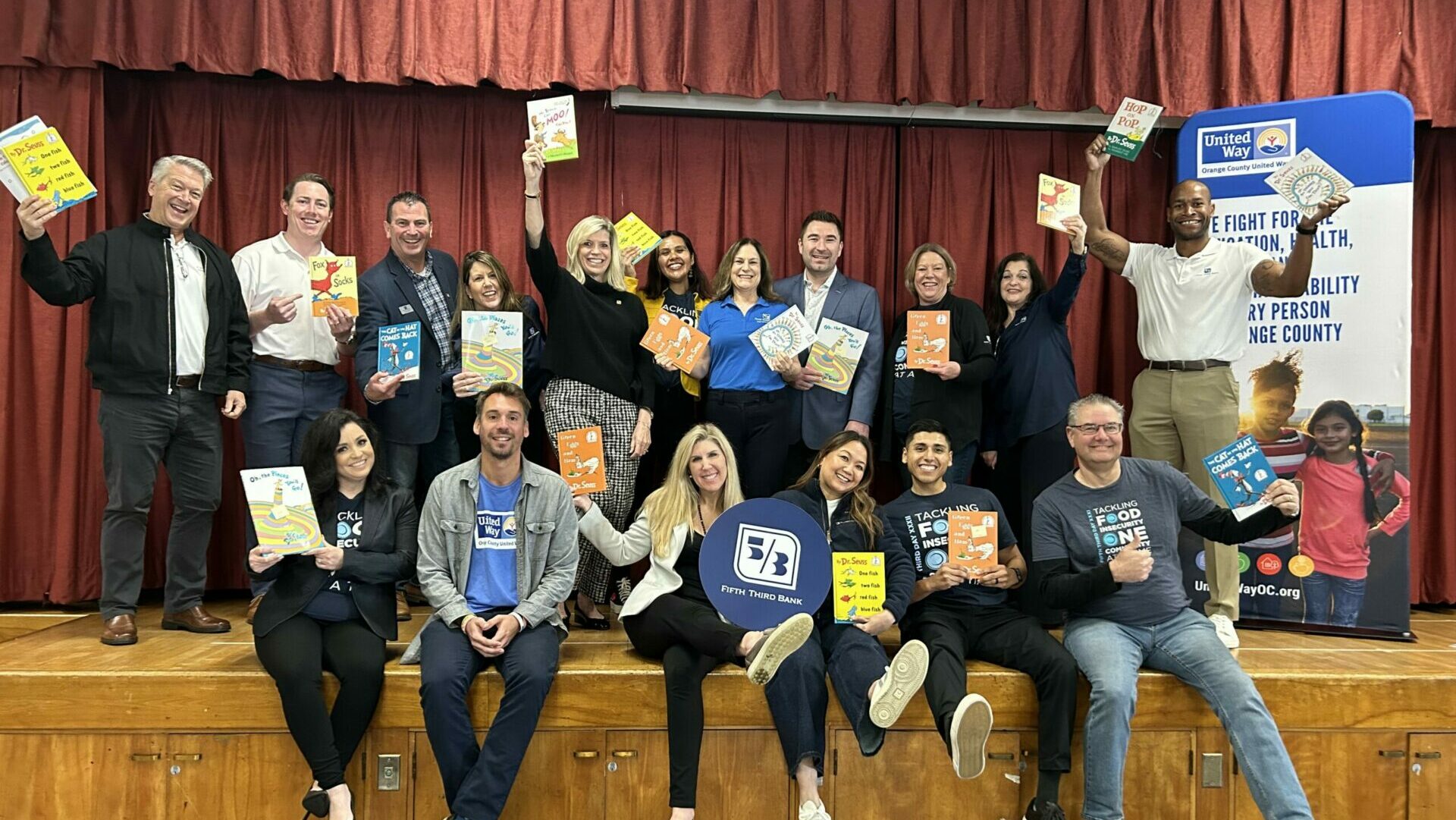 a group of adults on a school auditorium stage holding children's books aloft