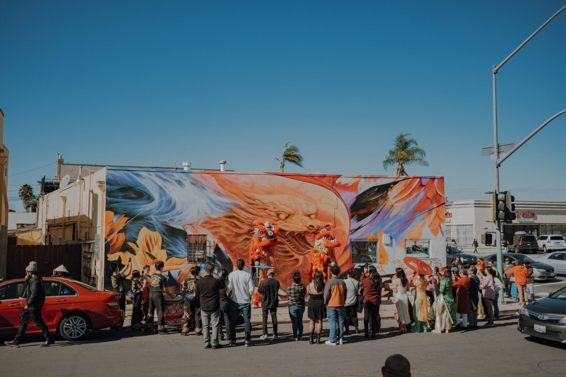 A group outside racing a blue, red, and while mural. In front of the crowd are performing Chinese Lion dancers. The sky is a bright blue