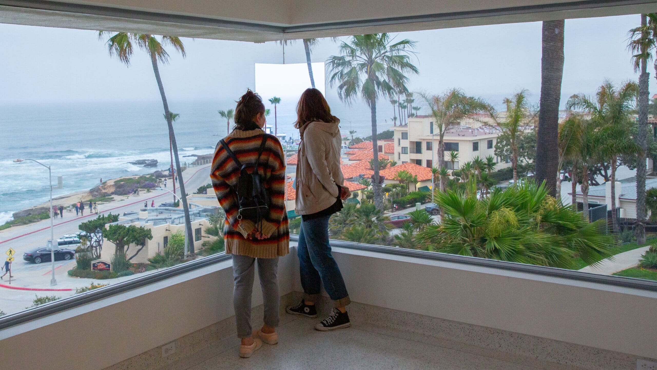 Two museum visitors standing and looking out a window that overlooks La Jolla and the Pacific Ocean.