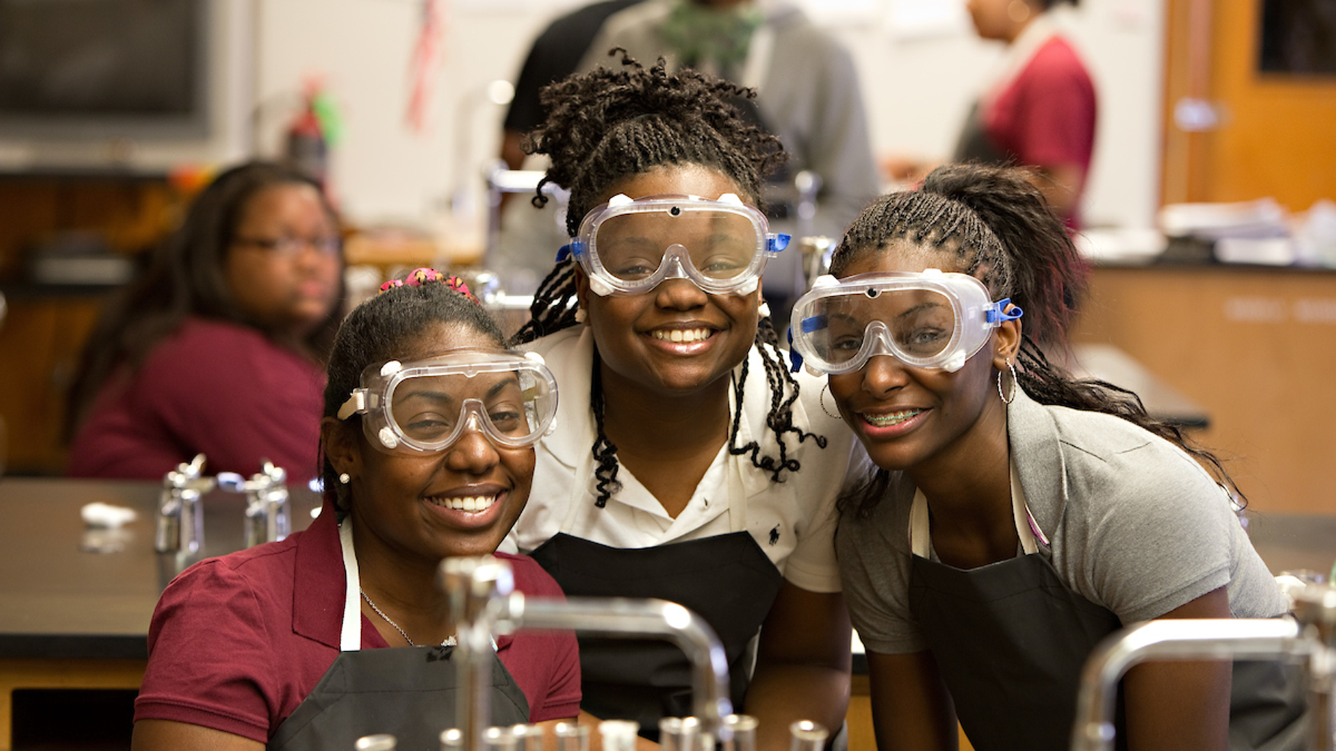Three students in science class wearing googles and smiling at camera