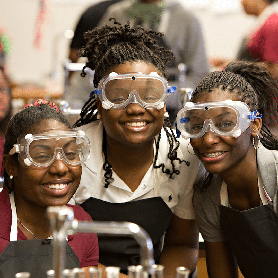 Three students in science class wearing googles and smiling at camera