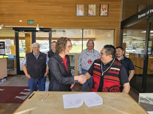 A group of people are gathered inside, two people shake hands in front of a signed document