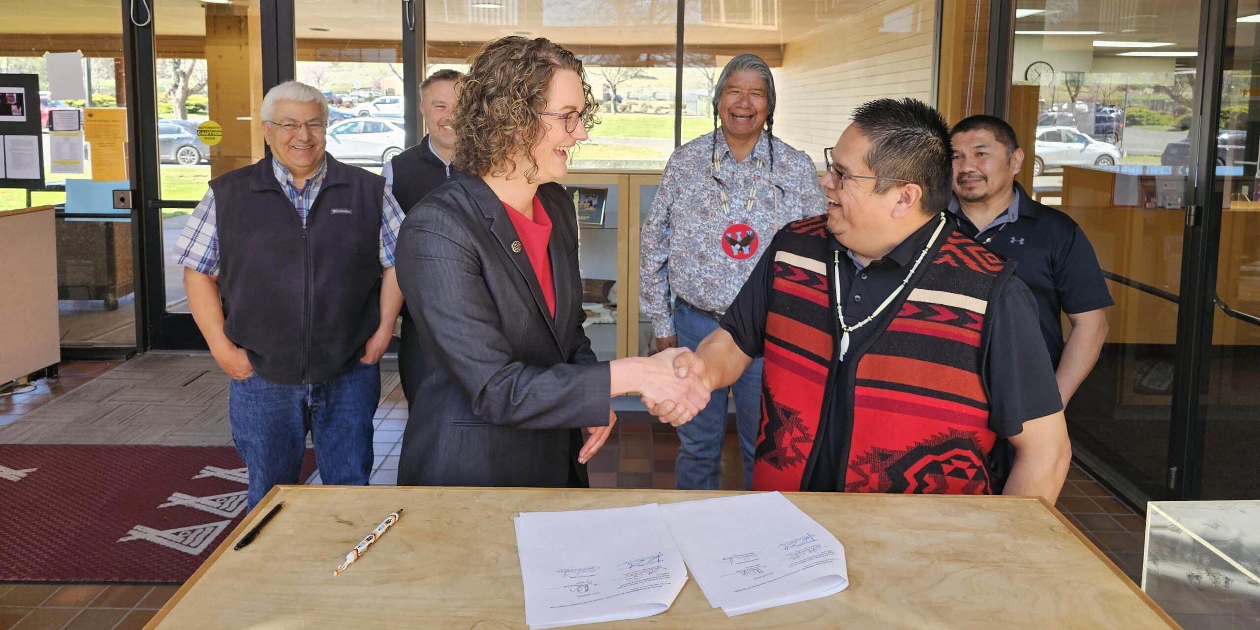 A group of people are gathered inside, two people shake hands in front of a signed document