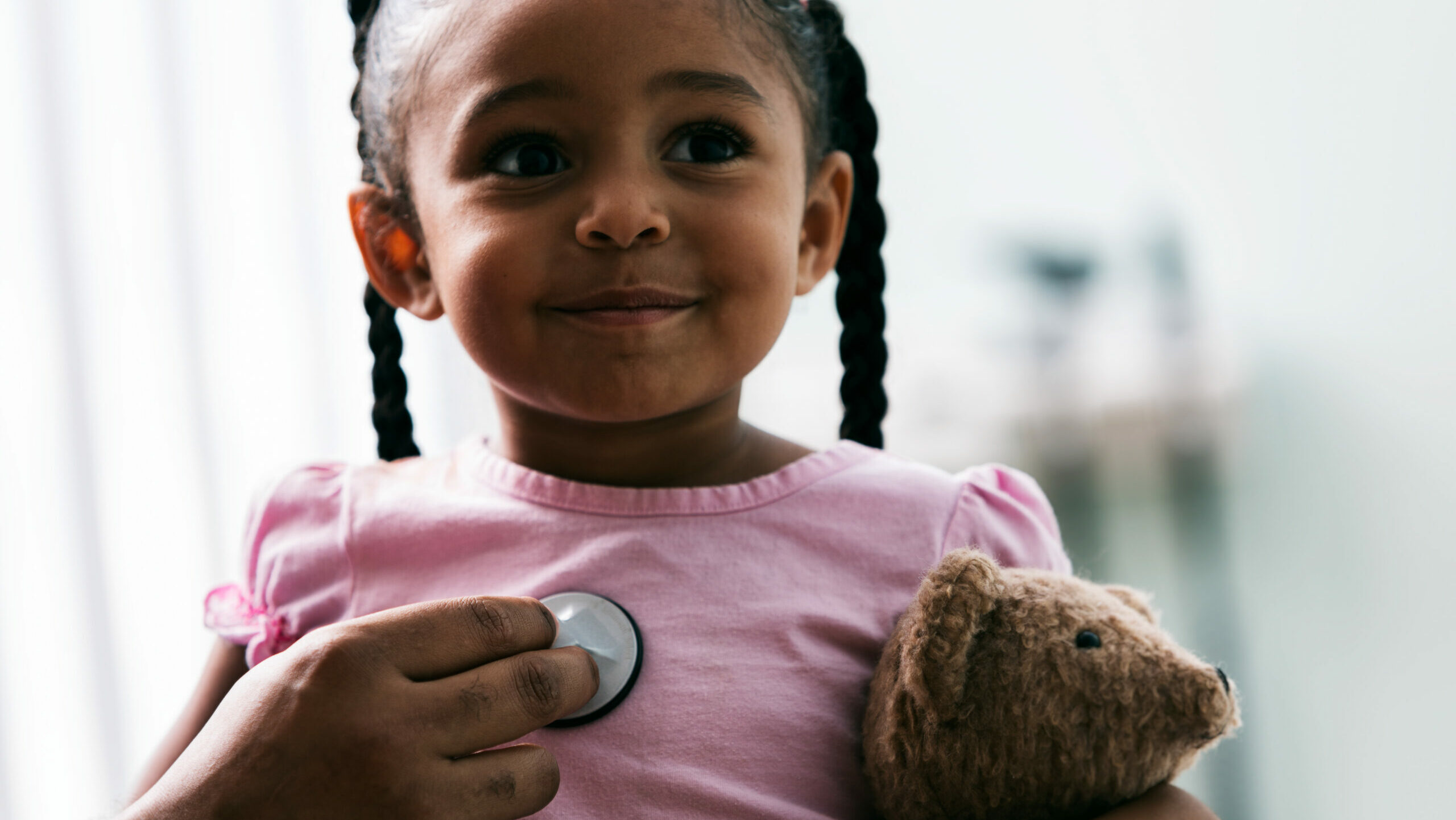 Girl holding a teddy bear while a doctor holds a stethoscope to her chest.